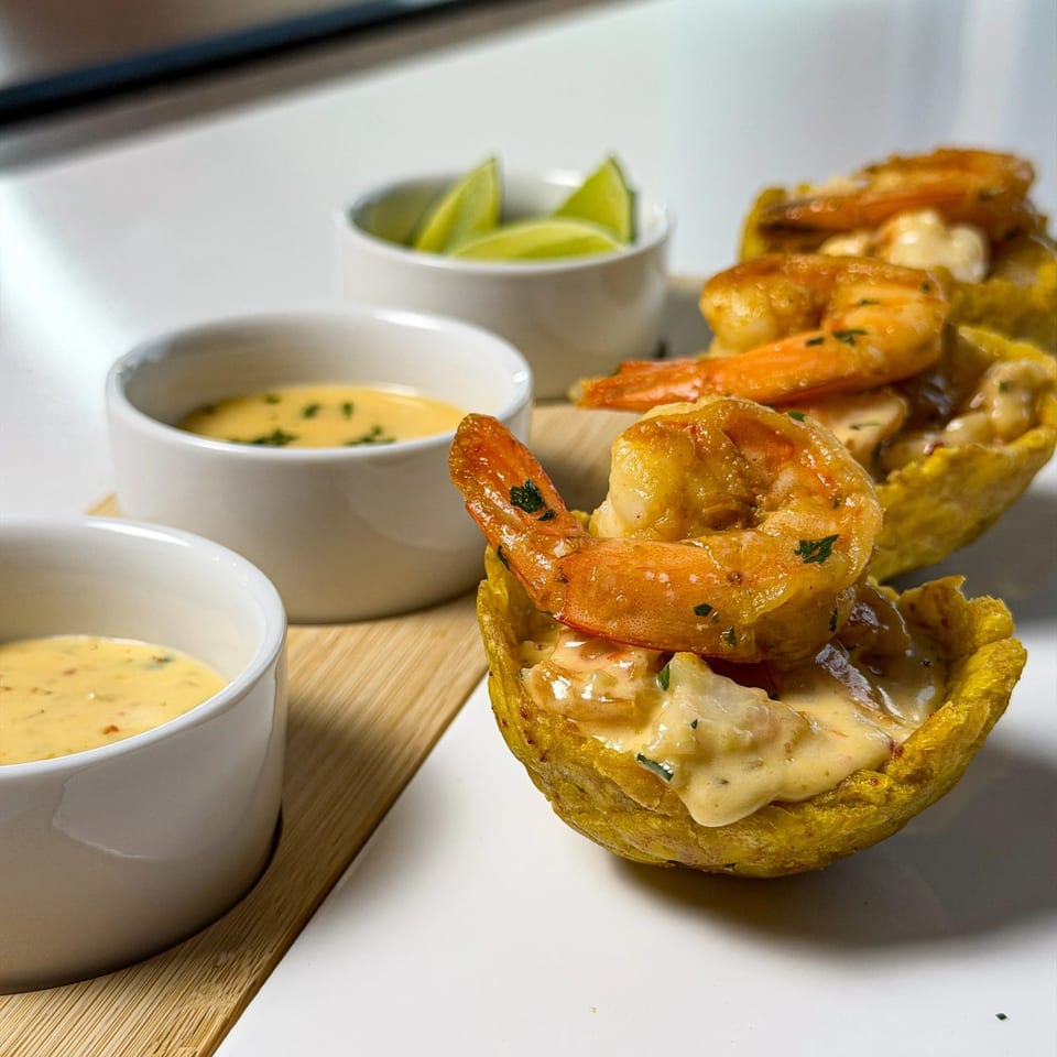 Shrimp tostones with creamy sauce and lime wedges on a white plate with a wooden board.