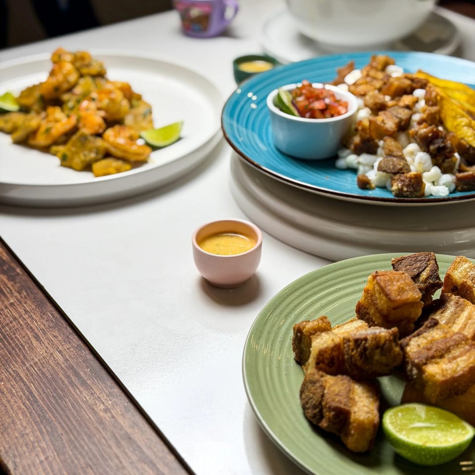 Three plates of Latin American food, including fried pork, shrimp, and plantains, on a white table.